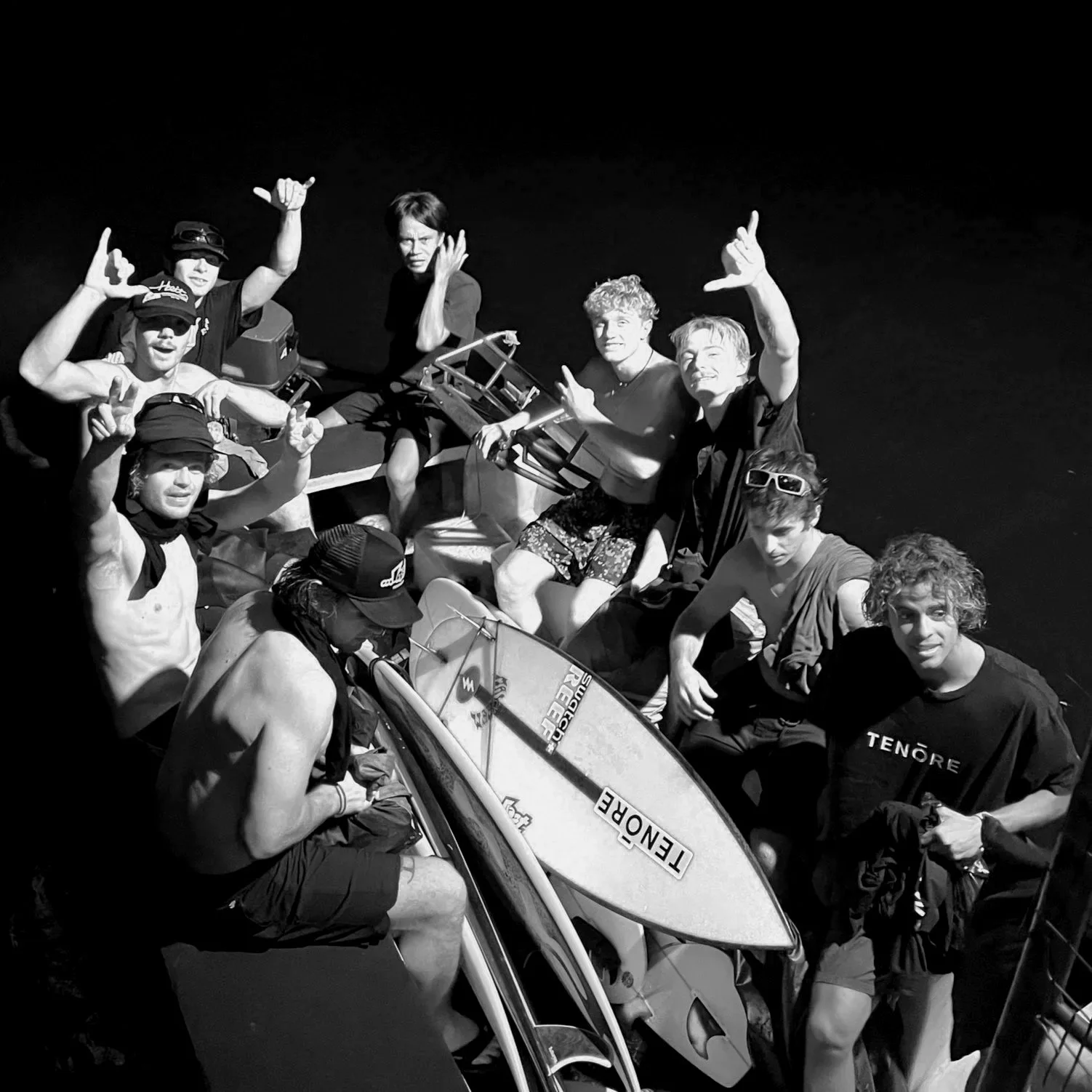 Group of young people on a boat at night, some making hand gestures, with surfboards on the boat, smiling and posing for the photo