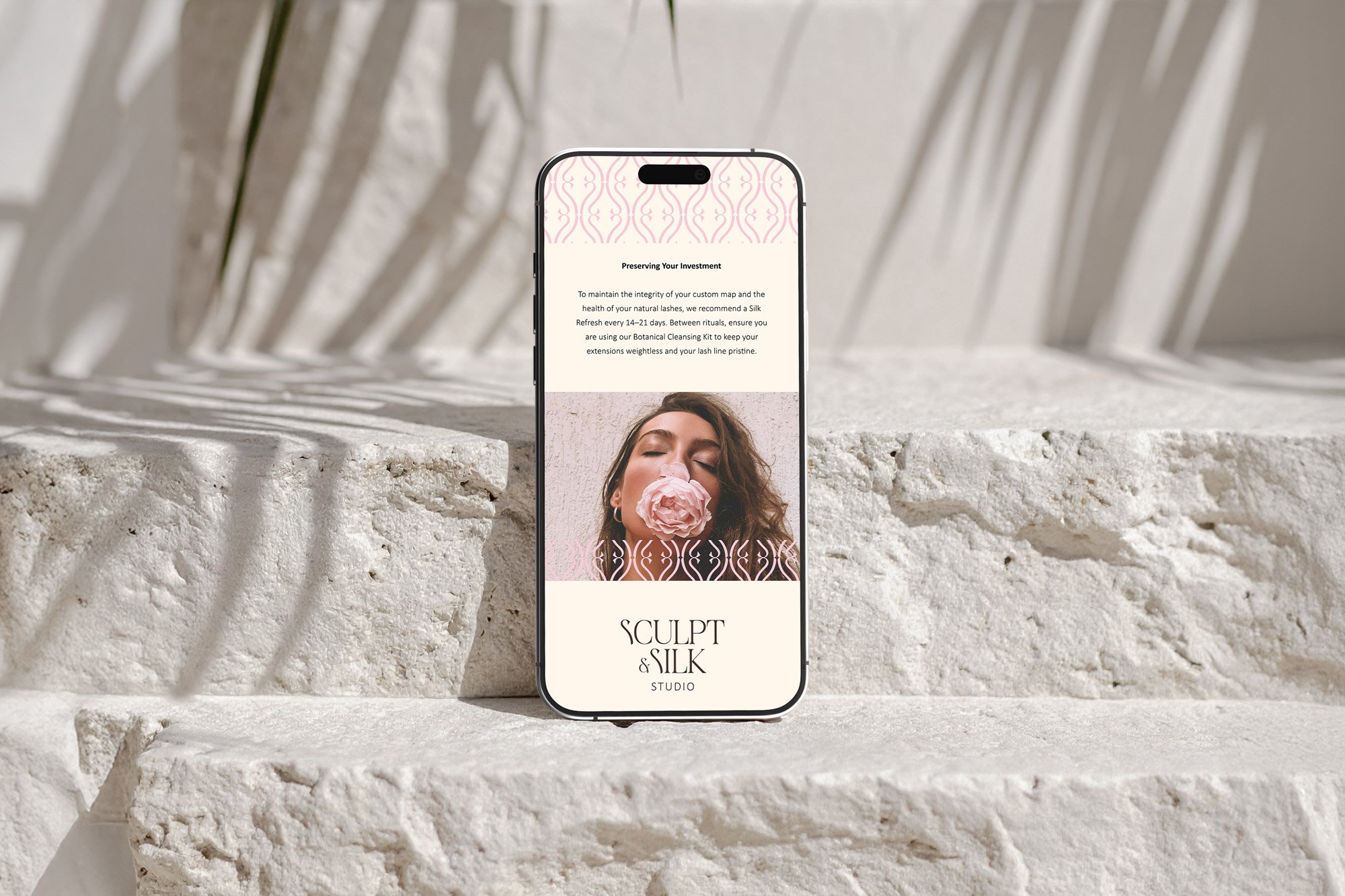 Smartphone with a website for 'Sculpt & Silk Studio' opened, displaying a woman with closed eyes holding a pink flower near her nose, against a textured, light-colored background.