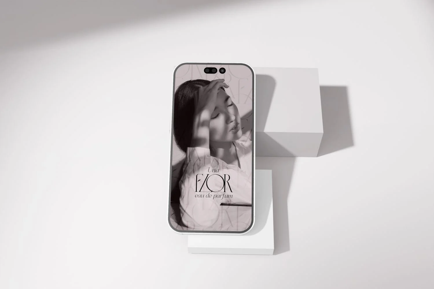 A smartphone displays a black and white photo of a woman with her eyes closed, touching her forehead. The phone is placed on a white pedestal against a minimalist white background. The screen shows the text 'Una FLOR eau de parfum'.