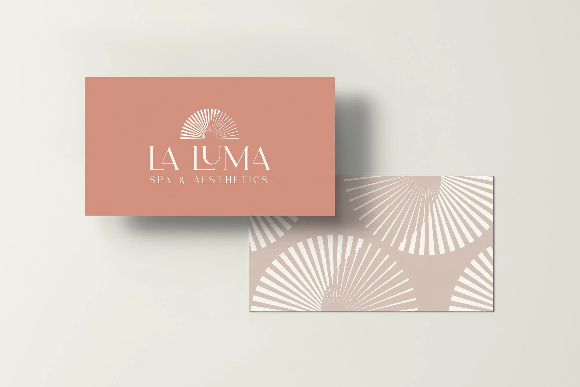 Business card for La Luma Spa & Aesthetics with a pink background, white text, and a sunburst logo; the back of the card has a light pink background with a pattern of white sunburst designs.