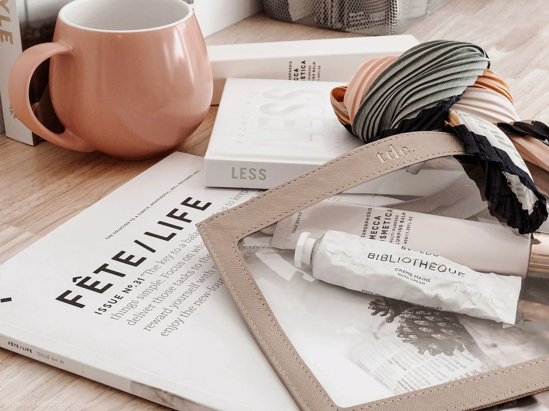 A pink mug, a magazine titled 'Fête Life', a copy of 'Less' by Rachel Cusk, a tube of hand cream labeled 'Bibliotheque', and a makeup brush wrapped in a black, white, and peach striped headband, all arranged on a wooden surface.