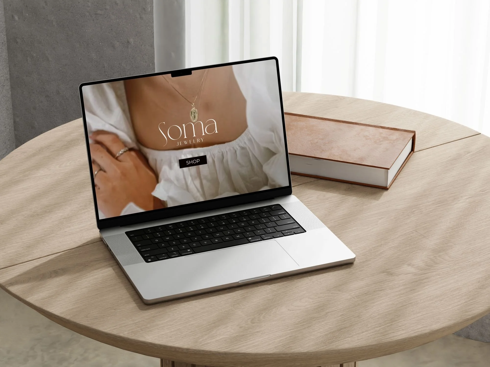 Laptop on a wooden table displaying a jewelry website called Soma Jewelry, with a model wearing a necklace and rings, and a closed book beside it.