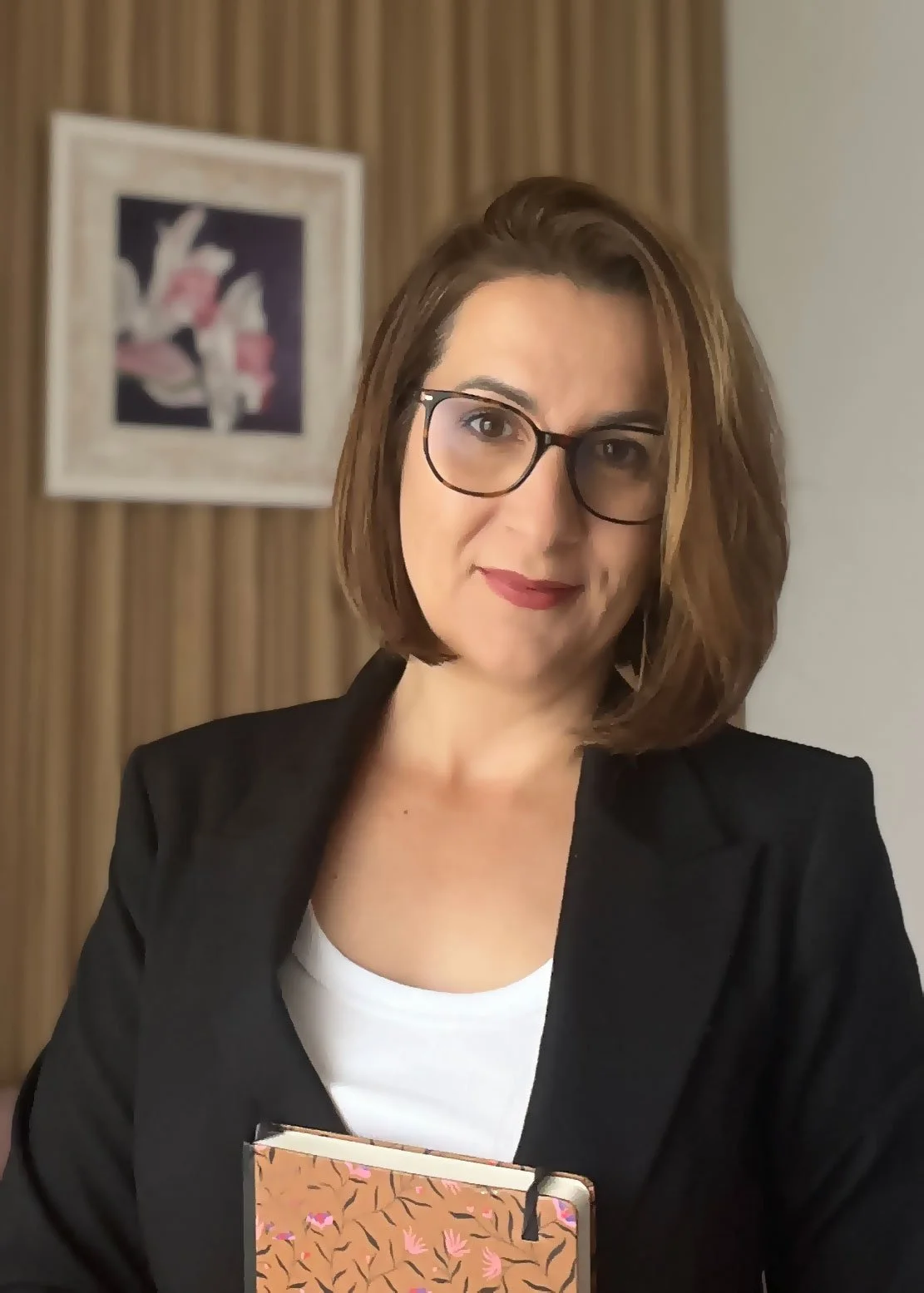 A woman with shoulder-length brown hair, glasses, and red lipstick, dressed in a black blazer and white top, holding a patterned notebook, standing in front of a framed artwork and wooden wall paneling.