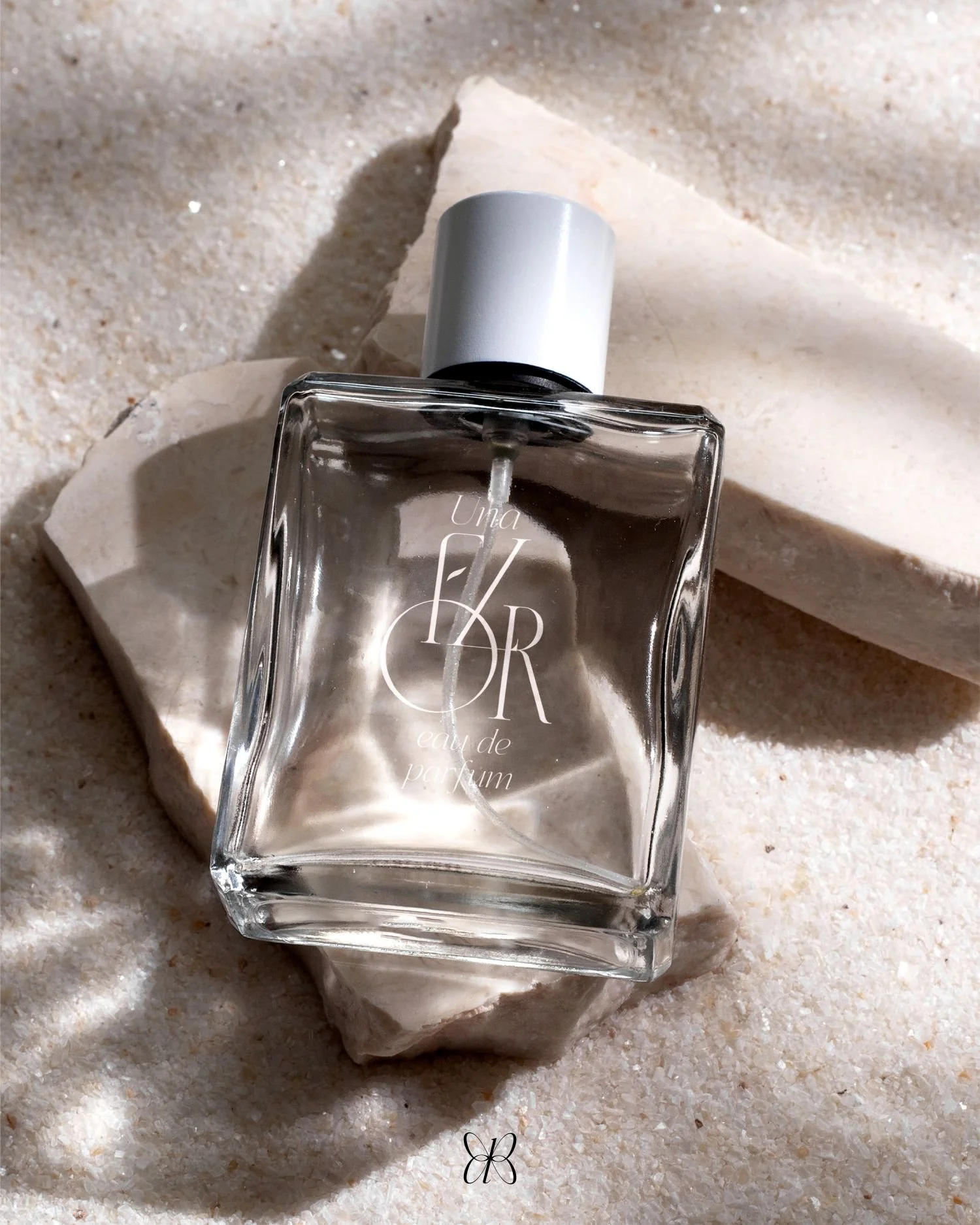 A square glass perfume bottle labeled 'Uno Cero' with a silver cap resting on a white textured stone on a beige sand surface with another larger stone in the background.