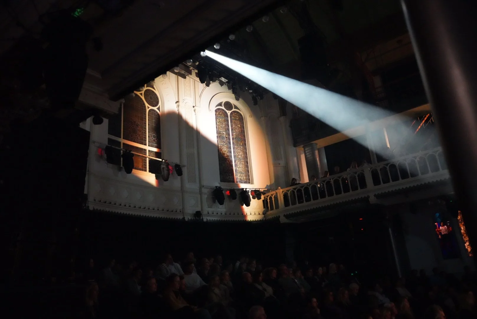 Interieur van een concertzaal met glas-in-loodramen en een schijnwerper die het publiek verlicht.