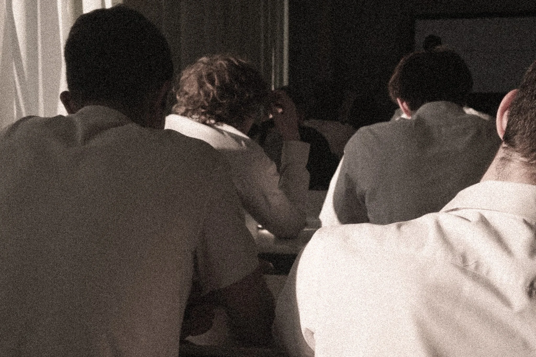 A group of people sitting closely together in a dimly lit room, with their backs to the camera, possibly attending a presentation or seminar.