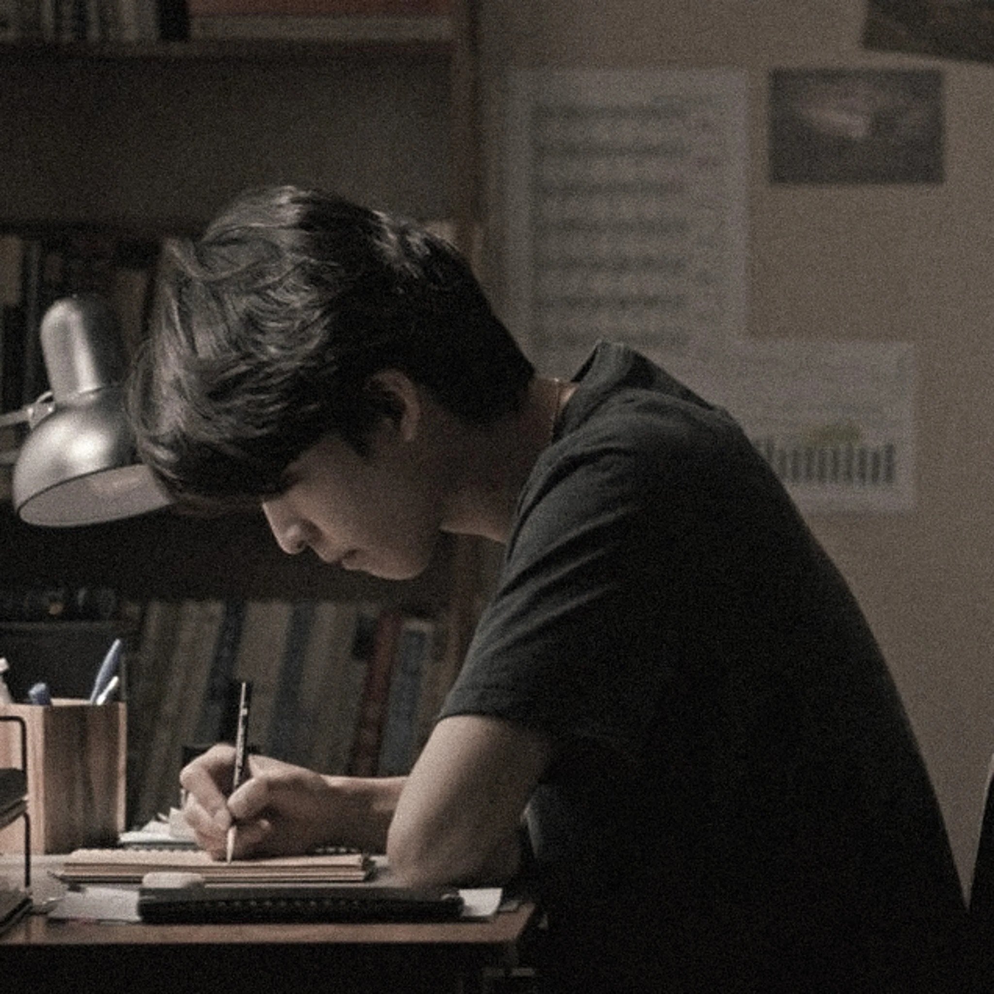 A person with long hair sitting on the floor, hunched over, possibly reading or writing, in a dimly lit room with shelves and boxes in the background.