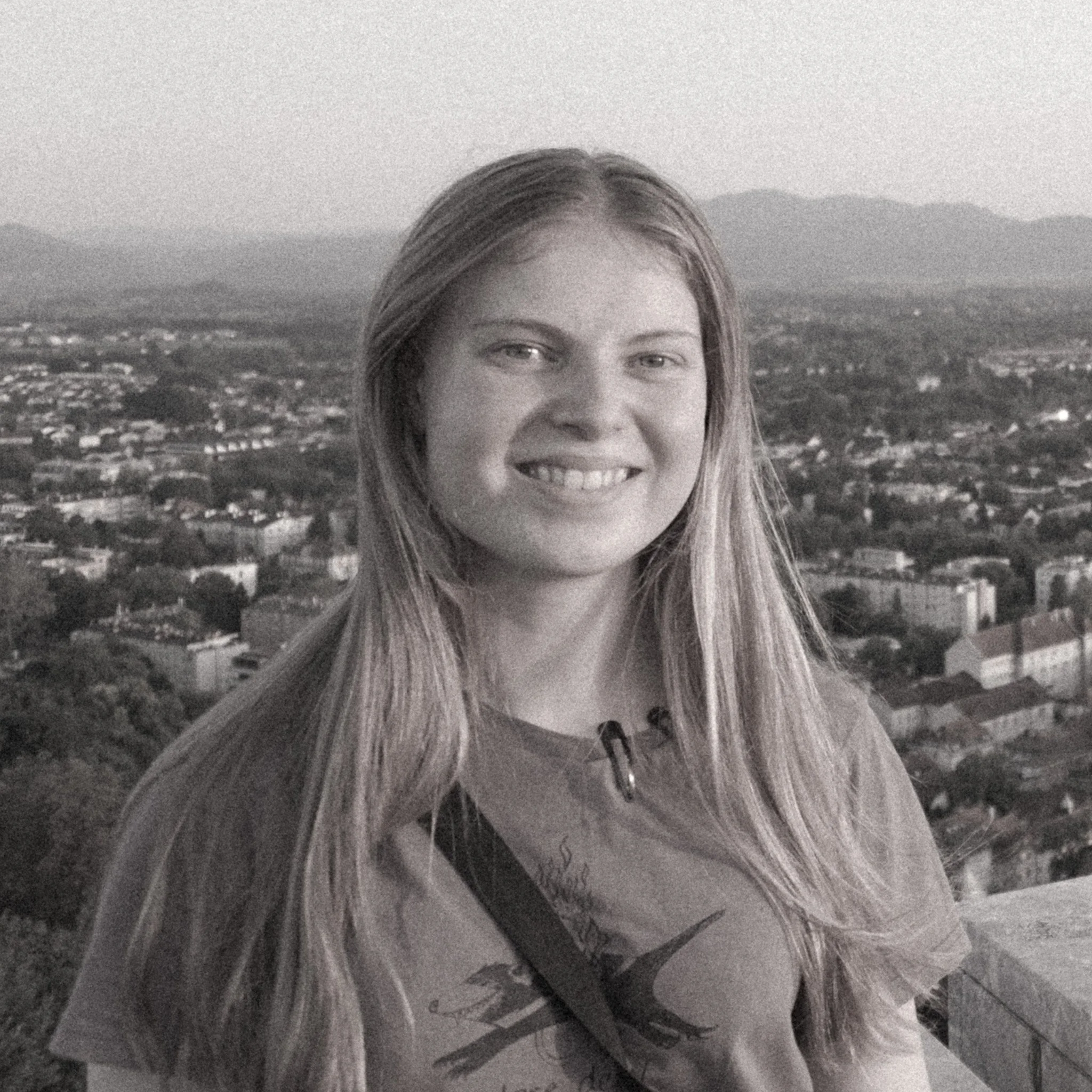 A woman with long hair smiling at the camera, with a landscape of mountains and a city in the background, in black and white.