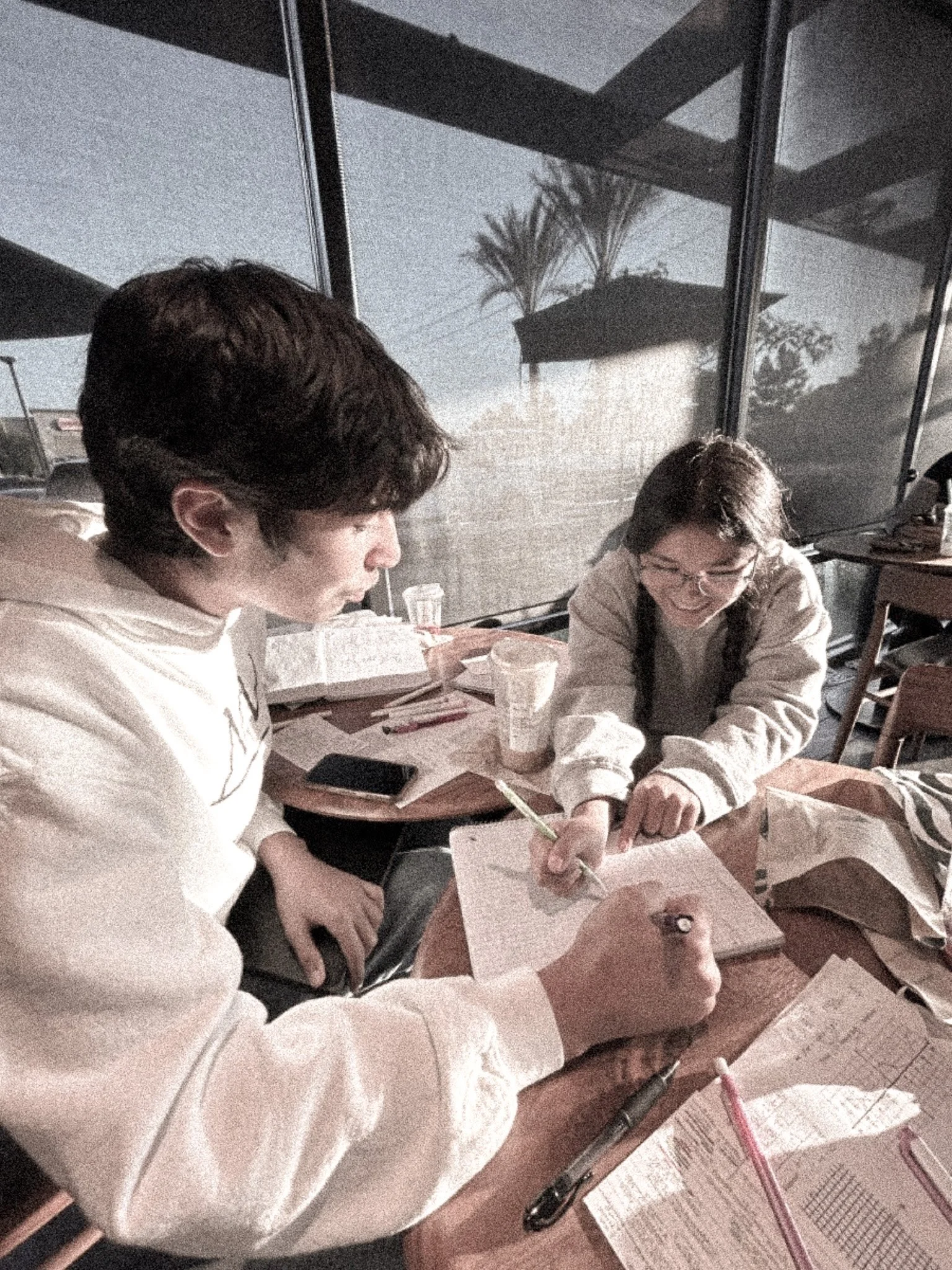 Two people, a man and a woman, are sitting at a wooden table, working on papers and writing with pens in a room with large window reflections of plants and trees outside.