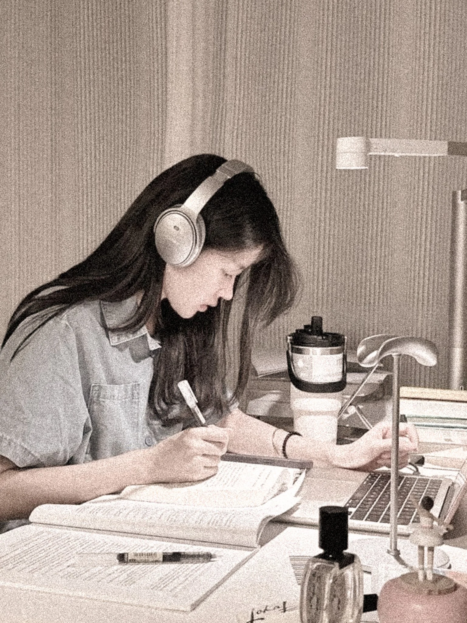 A young woman with long dark hair, wearing headphones, writing notes at a desk in a workspace, with a laptop, notebooks, and a water bottle nearby.