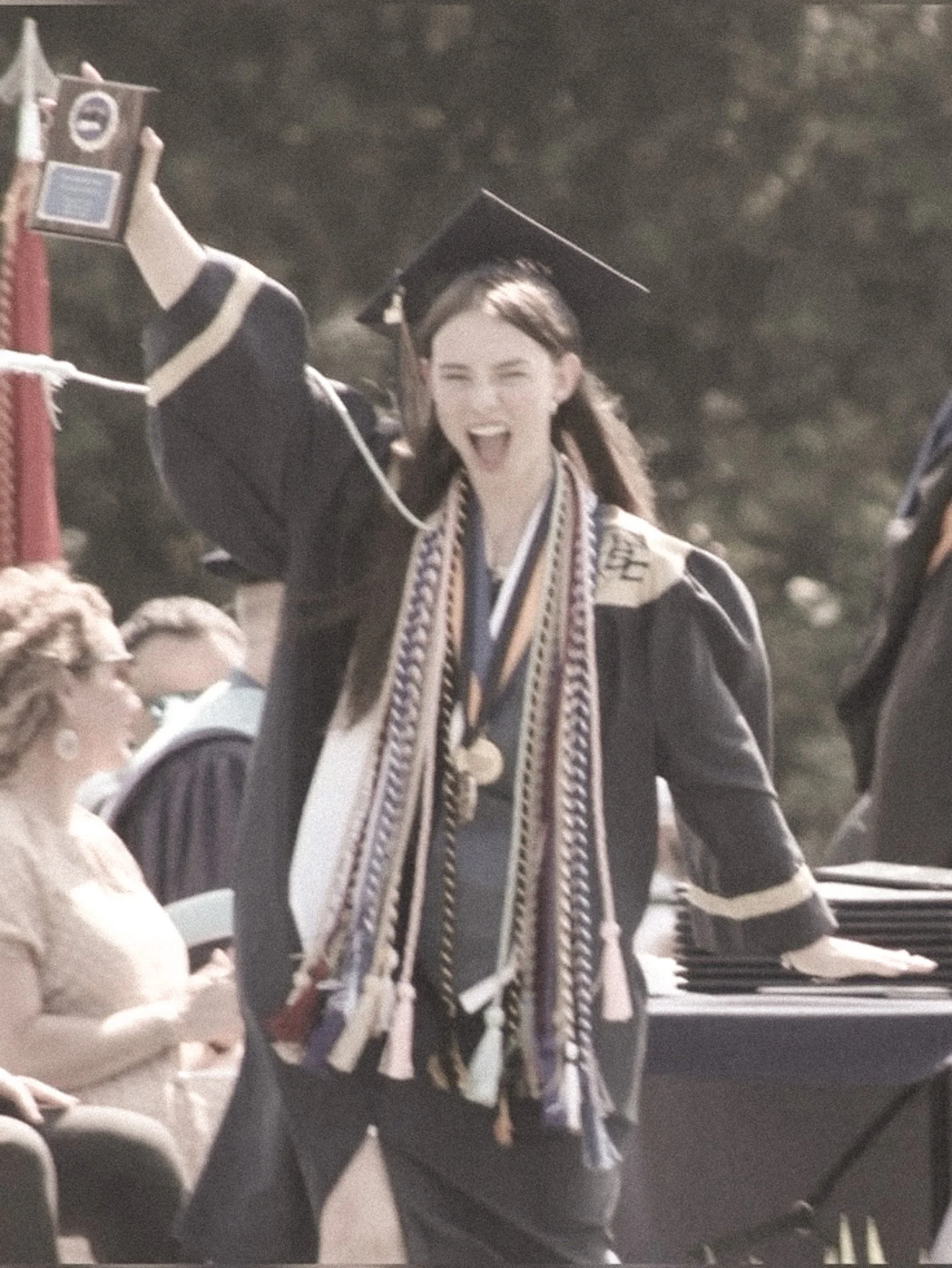 A woman in graduation cap and gown celebrating with a joyful expression, with tassels and cords around her neck, at what appears to be a graduation ceremony.