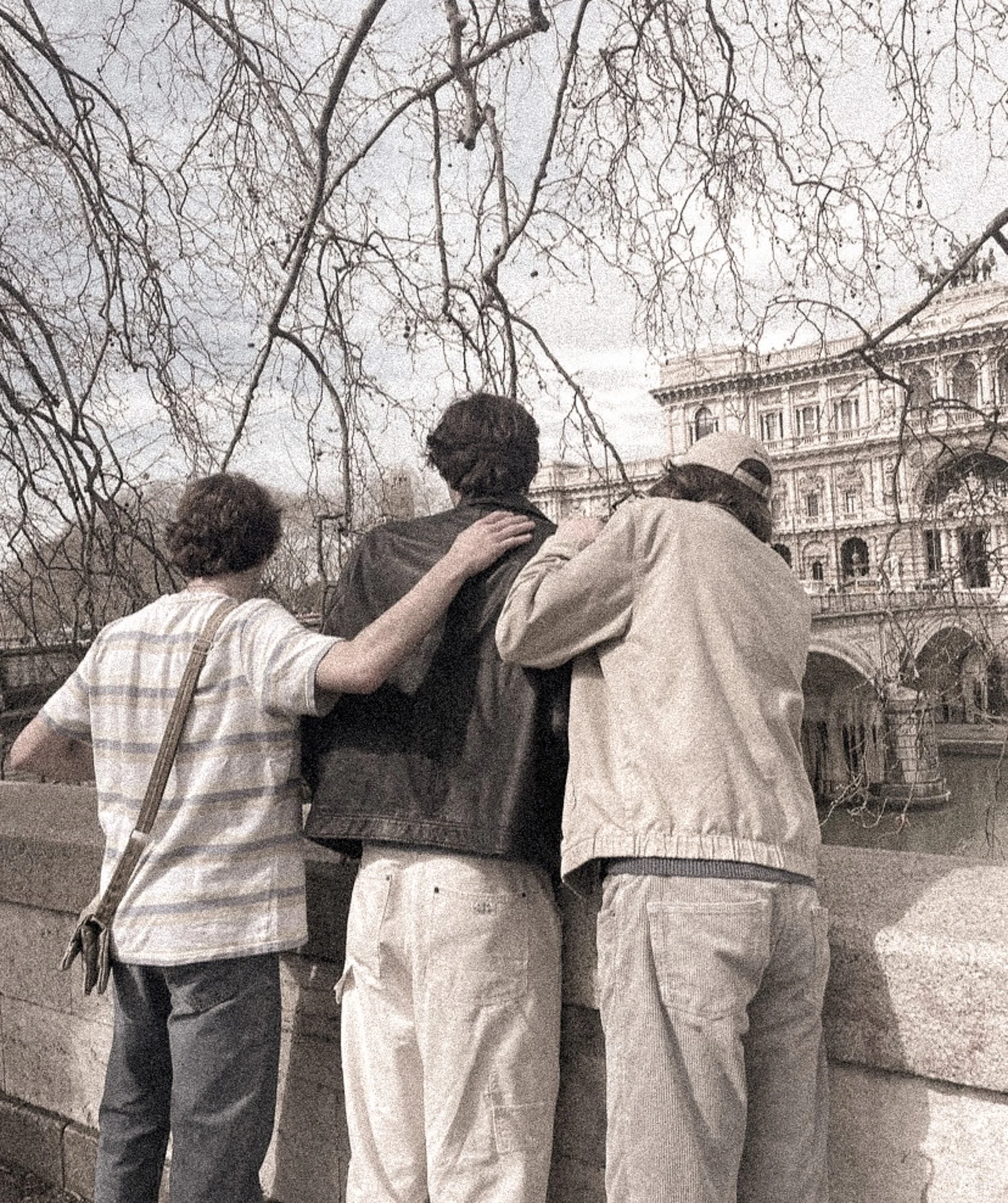 A historical black-and-white photo of three people with their arms around each other's shoulders, standing outside in front of a large, ornate building with multiple windows and architectural details, under leafless trees.