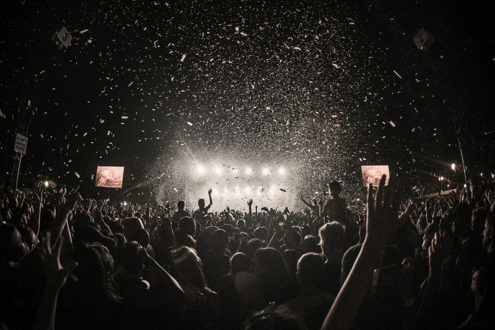 Crowd at a concert or event with confetti falling from above and stage lights in the background.