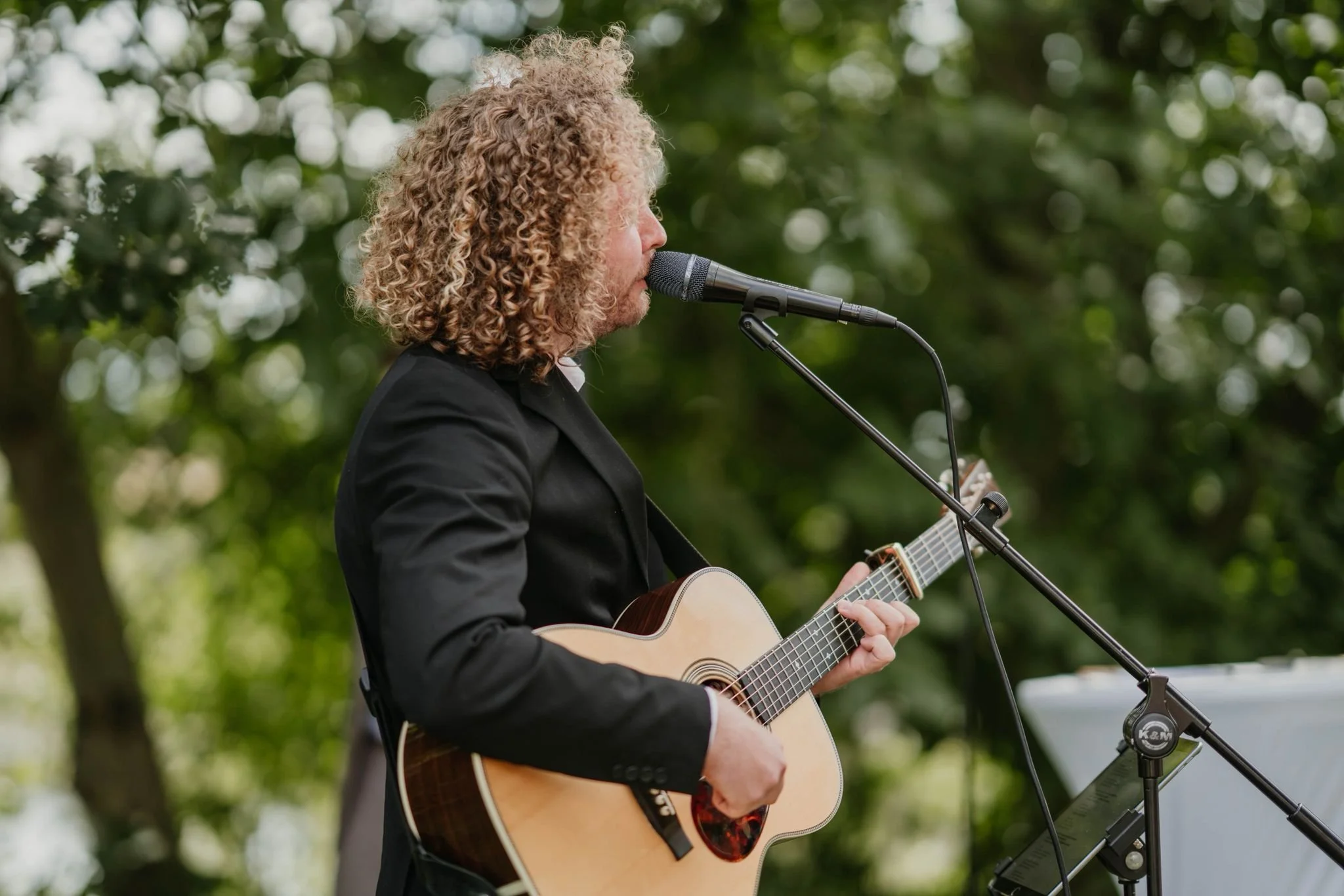 Ein Mann mit lockigem Haar spielt Gitarre und singt in der Natur vor einem Mikrofon.