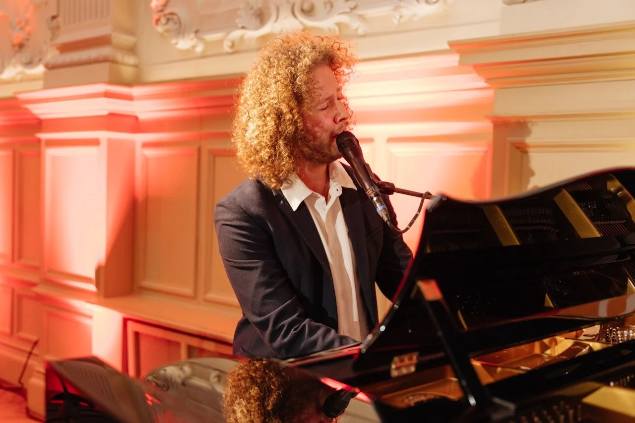 Ein Mann mit lockigem, blondem Haar spielt Klavier und singt in einem Saal mit eleganter Innenarchitektur.