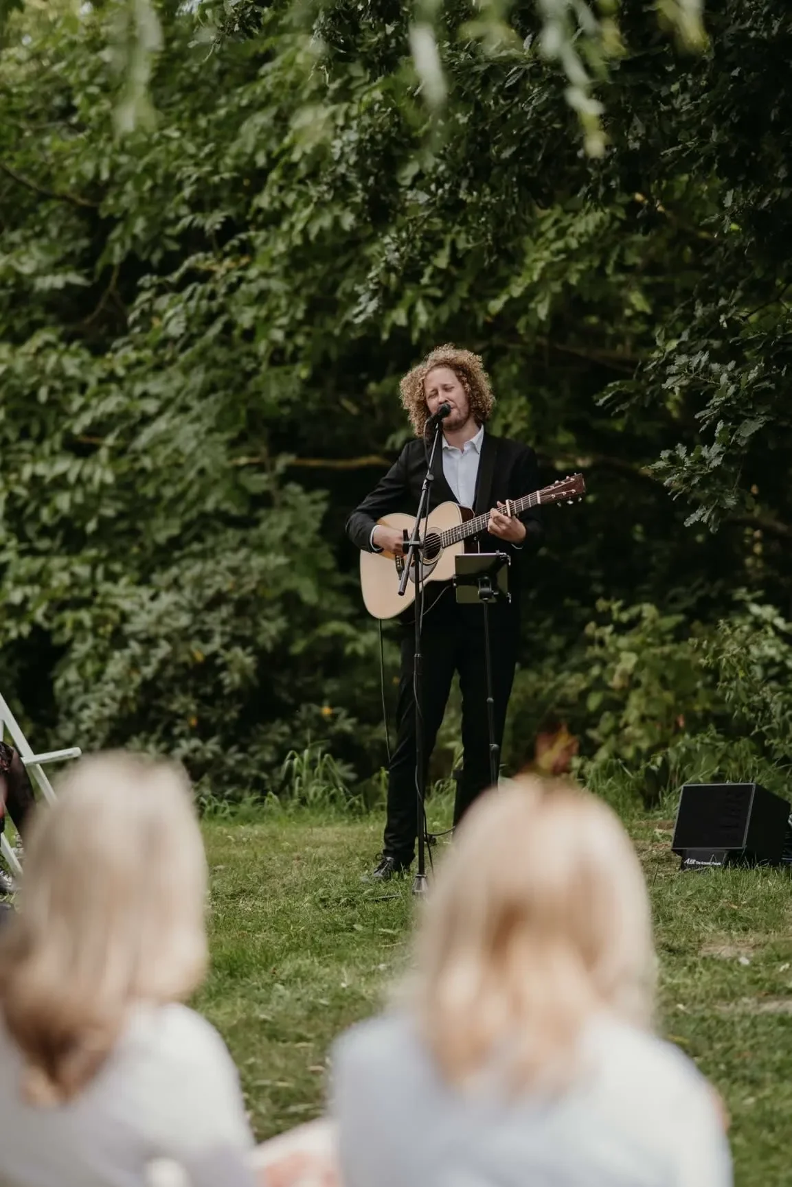Ein Mann mit lockigem Haar singt und spielt Gitarre bei einer Outdoor-Performance, während er vor einem Publikum steht, umgeben von grünen Bäumen.