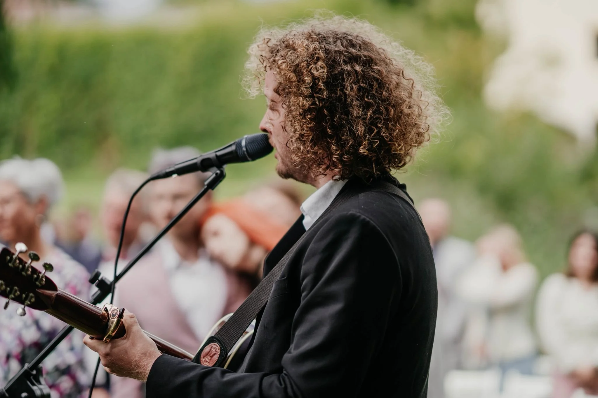 Ein Mann mit lockigem Haar spielt Gitarre und singt in einer Gruppe im Freien.