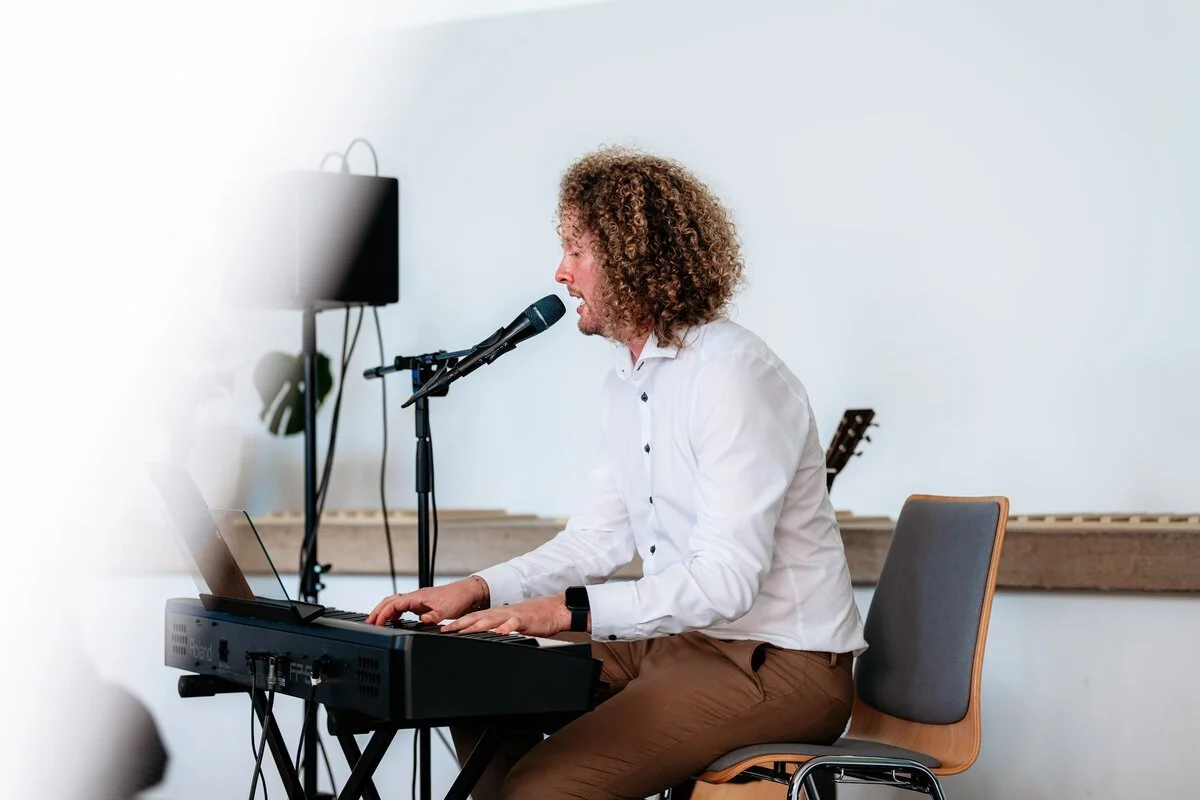 Mann spielt Klavier und singt in einem Raum, möglicherweise bei einer Aufnahme oder Aufführung, mit einem Mikrofon und einem Laptop.