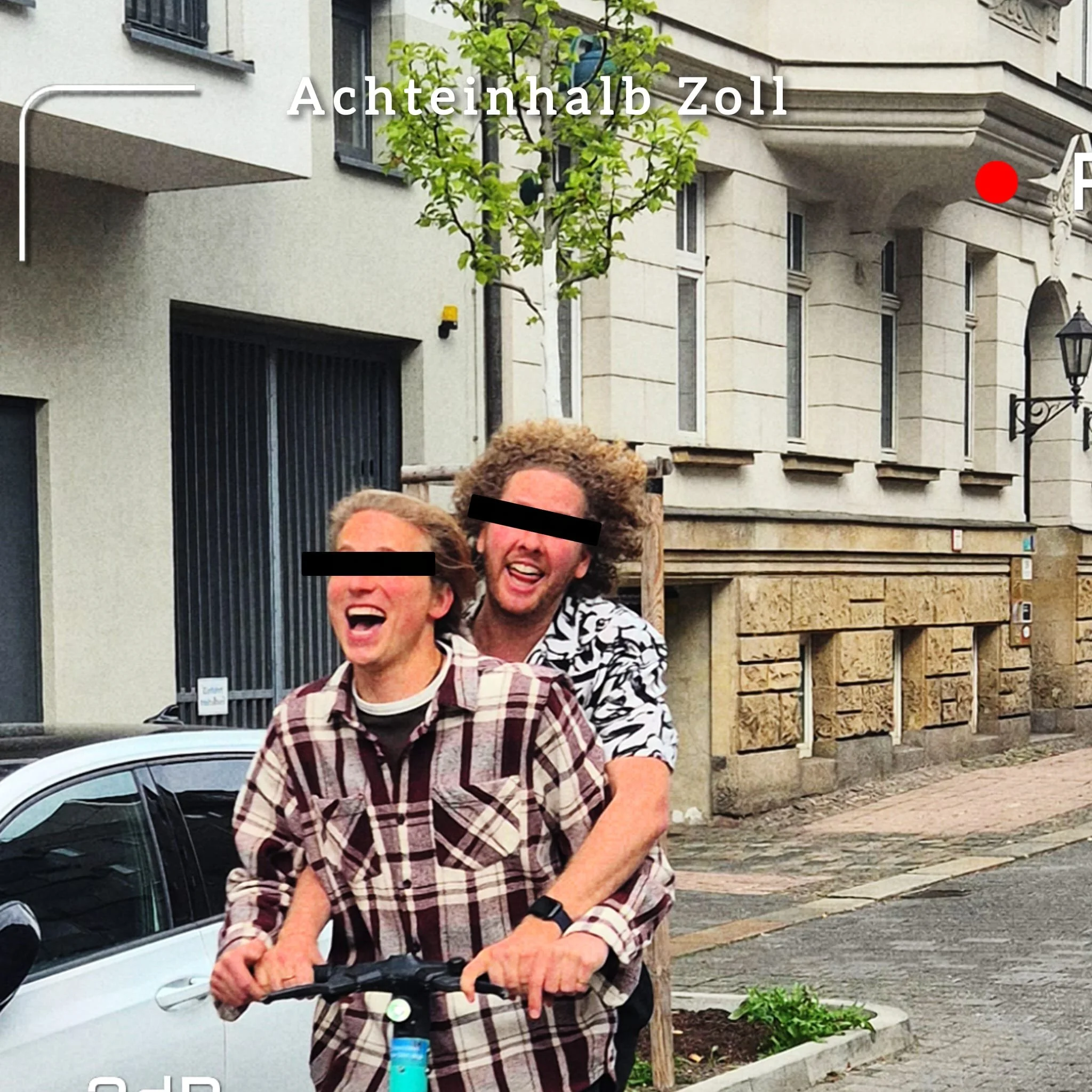 Zwei lachende junge Männer, einer mit braunen kurzen Haaren, der andere mit lockigem braunem Haar, stehen auf der Straße vor einem modernen Mehrfamilienhaus, in einer urbanen Umgebung, während sie ein Elektro-Faltrad halten.