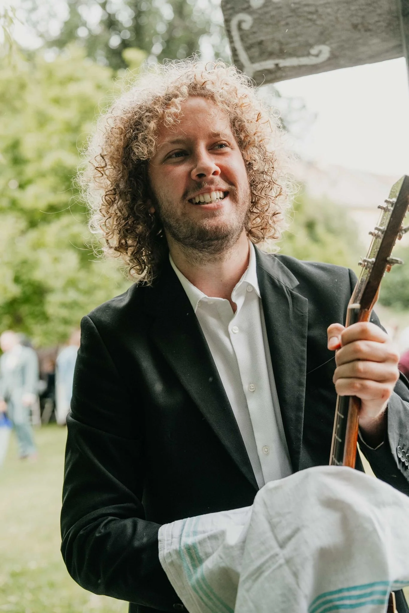 Ein Mann mit lockigem Haar trägt einen schwarzen Anzug und spielt Gitarre auf einer outdoor Veranstaltung, im Hintergrund sind Bäume und Leute zu sehen.