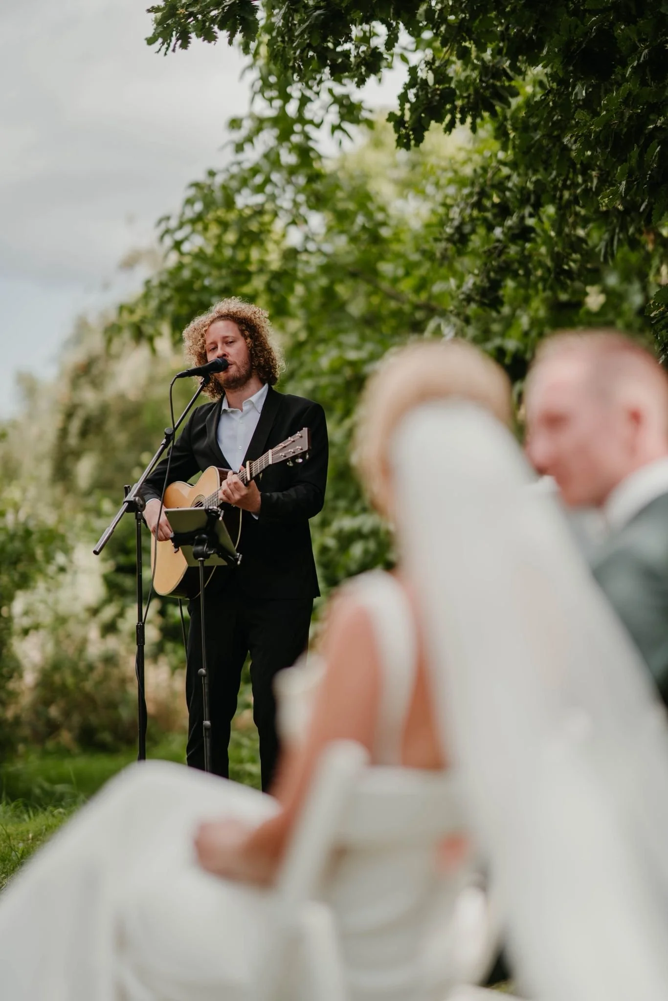 Mann spielt Gitarre und singt bei einer Veranstaltung im Freien, im Hintergrund sind Bäume und eine graue Wolkendecke, im Vordergrund sind ungeordnete, weiße, geschlossene Blumen bei einer Hochzeit zu sehen.