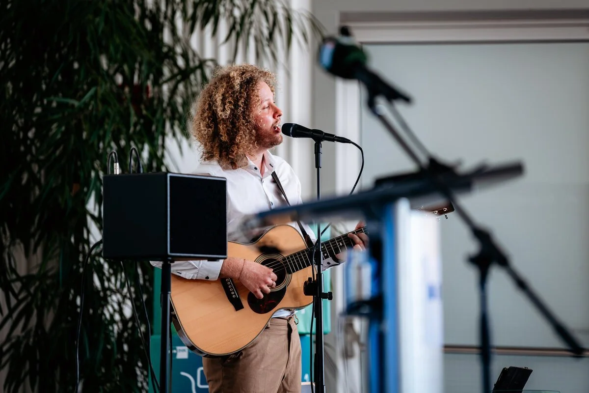 Ein Mann mit lockigem Haar spielt Gitarre und singt vor einem Mikrofon, während er bei einer Aufzeichnung oder einem Live-Auftritt ist, im Hintergrund Pflanzen.