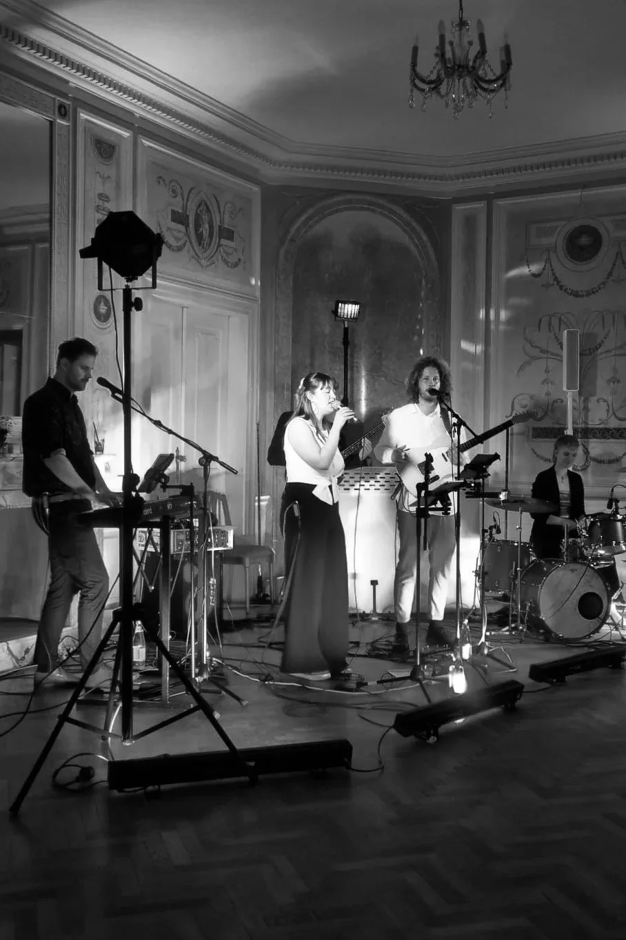Eine Musikband spielt in einem eleganten, historischen Raum mit aufwändiger Wanddekoration und Kronleuchter. Das Bild ist in Schwarz-Weiß aufgenommen.