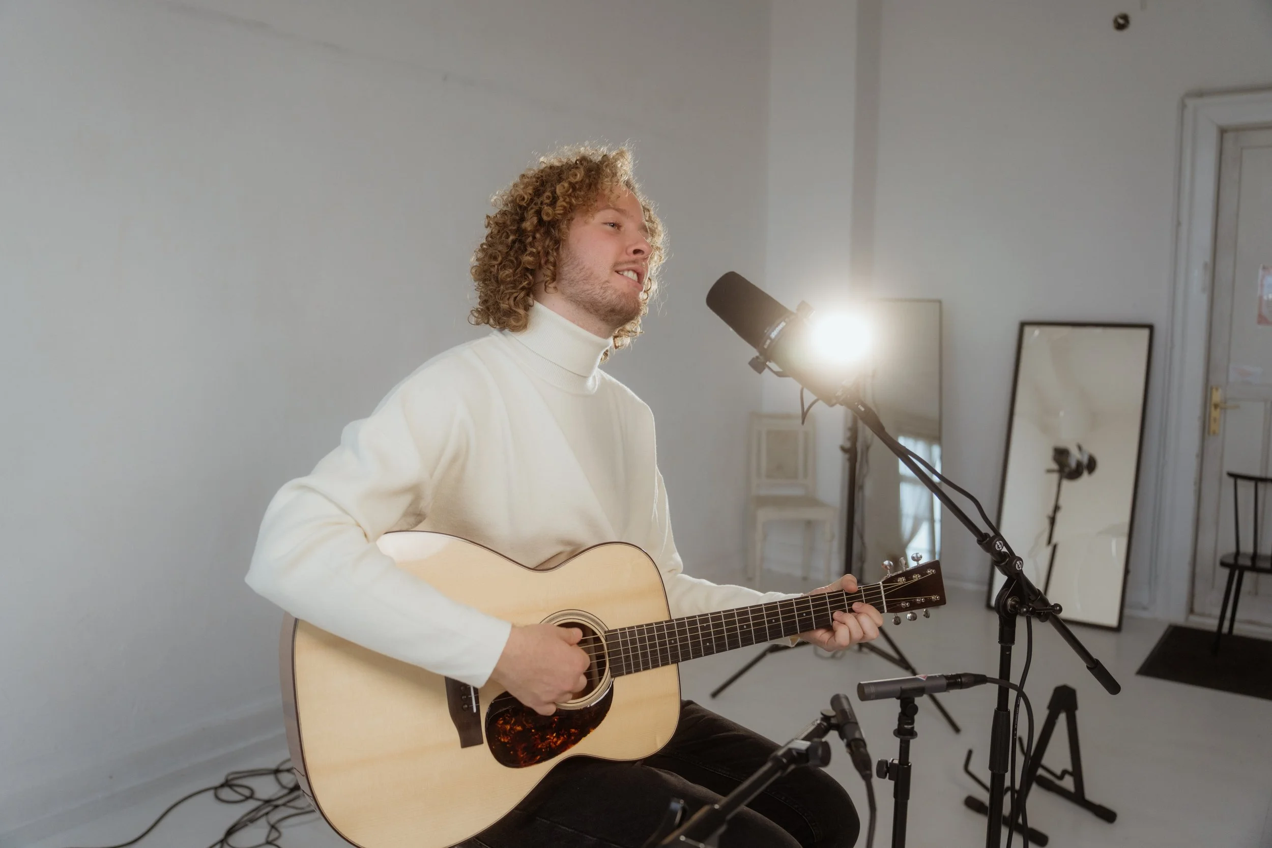 Ein junger Mann mit lockigem Haar spielt Gitarre und singt in einem Studio. Es gibt mehrere Mikrofone und Spiegel im Hintergrund.