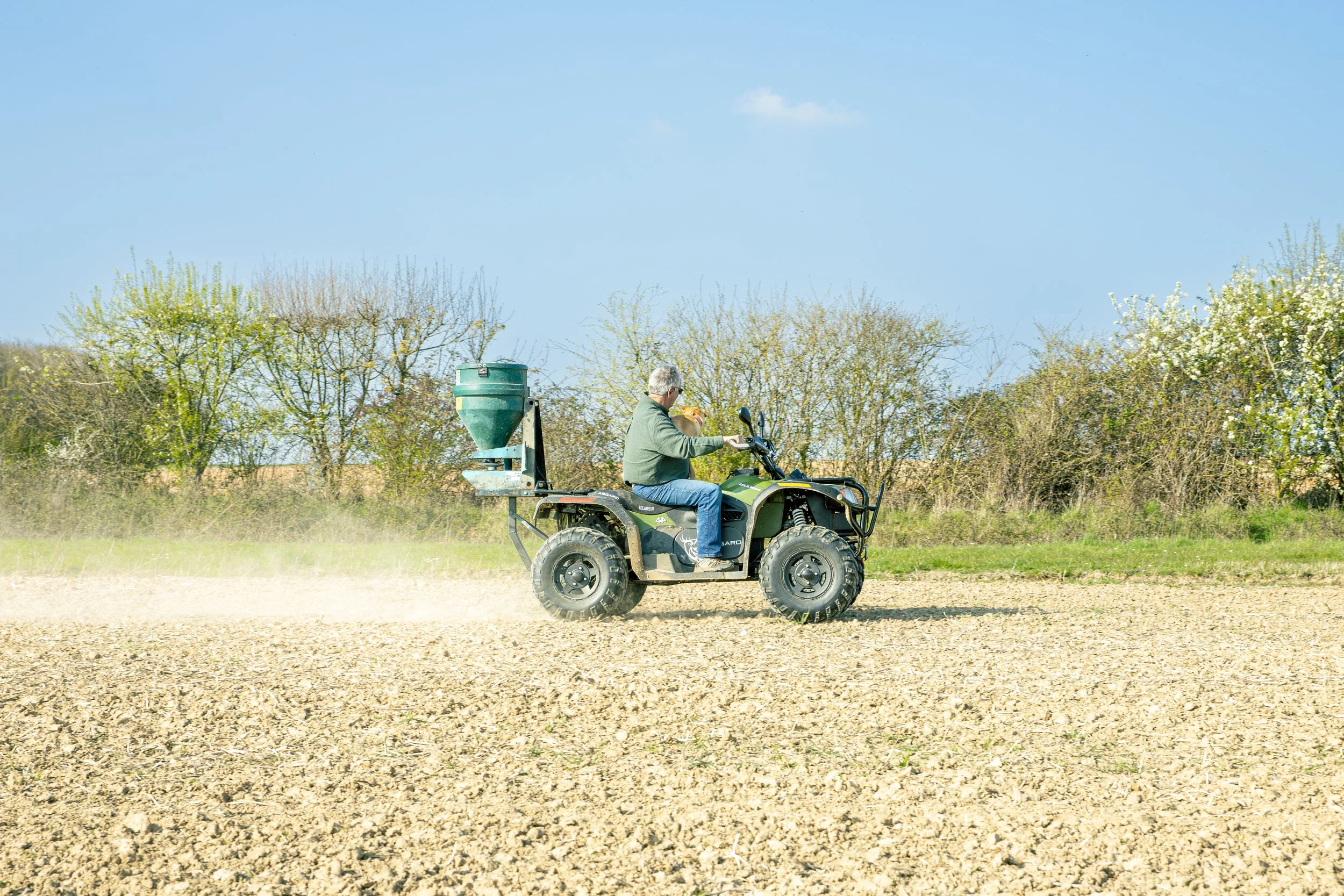Un homme conduit un quad dans un champ agricole avec des arbres en arrière-plan, durant une journée ensoleillée.
