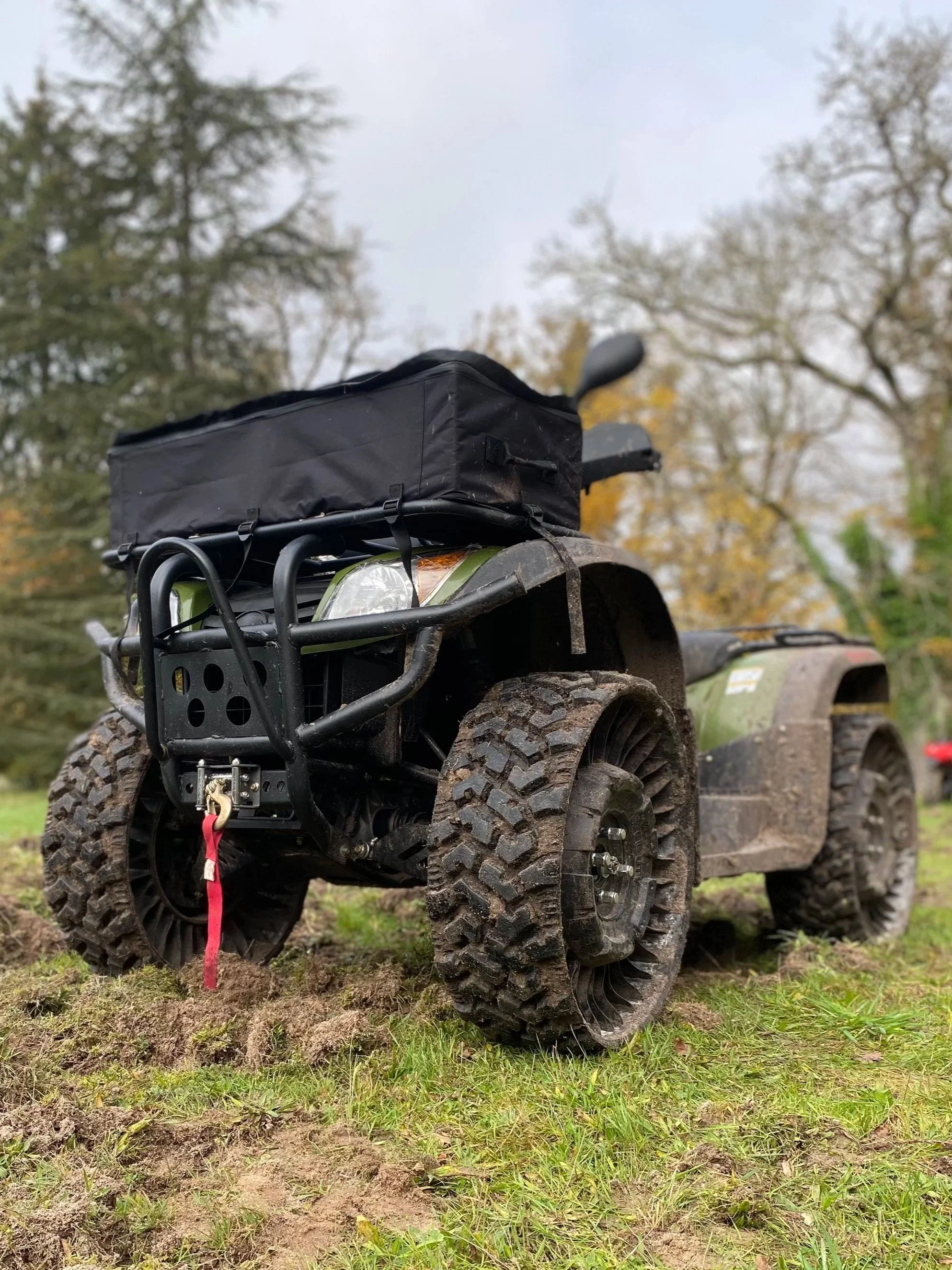Une moto tout-terrain avec des pneus très larges, un sac noir fixé à l'avant et un crochet rouge attaché à l'avant de la moto, sur un terrain herbeux avec des arbres en arrière-plan.