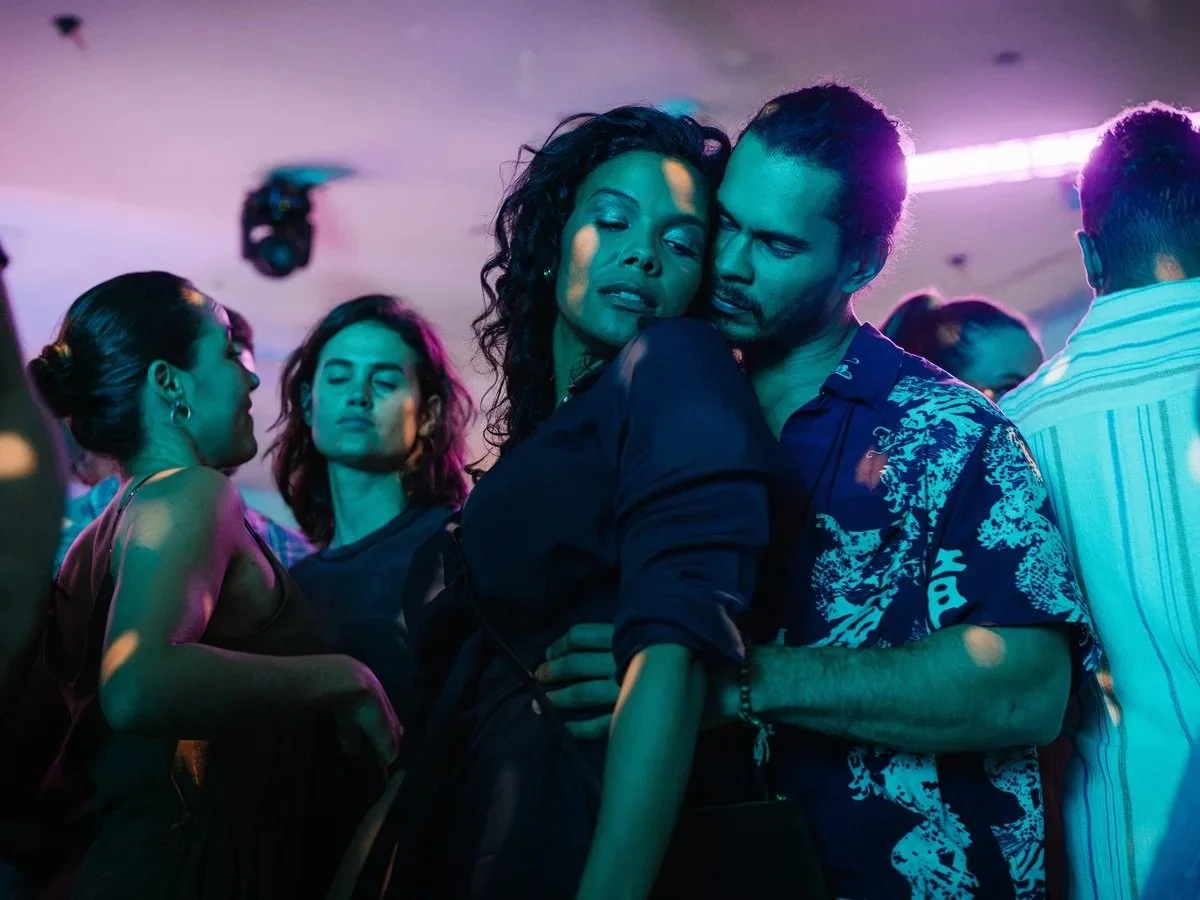 Couple dancing closely at a nightclub with colorful neon lighting, surrounded by other partygoers.