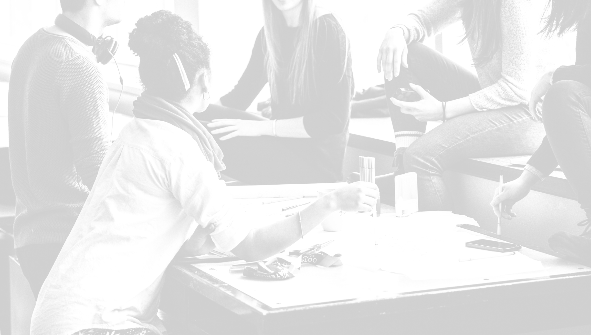 A group of people gathered around a table in a casual setting, some sitting and one leaning, engaging in conversation or an activity.