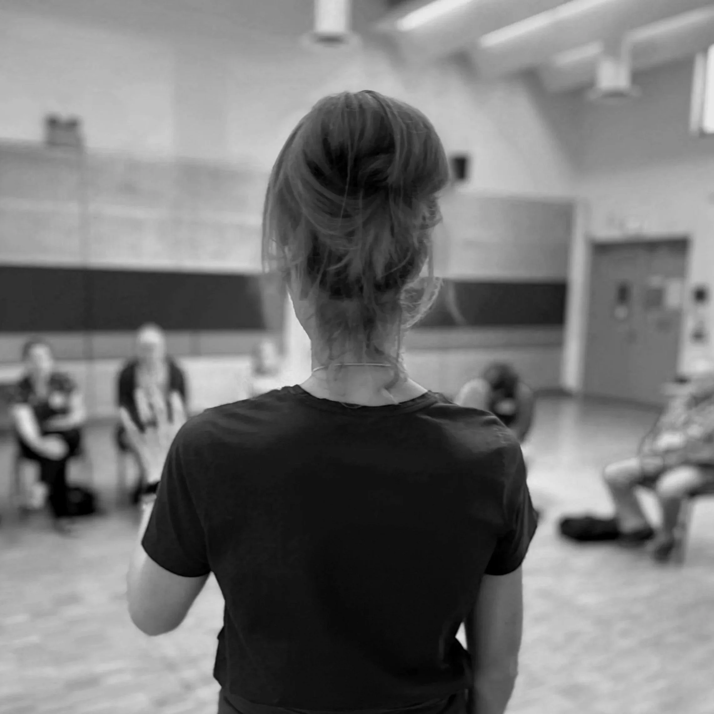 Back view of a woman with short hair in a black t-shirt standing in a room with a group seated on chairs, engaging in a discussion or class.
