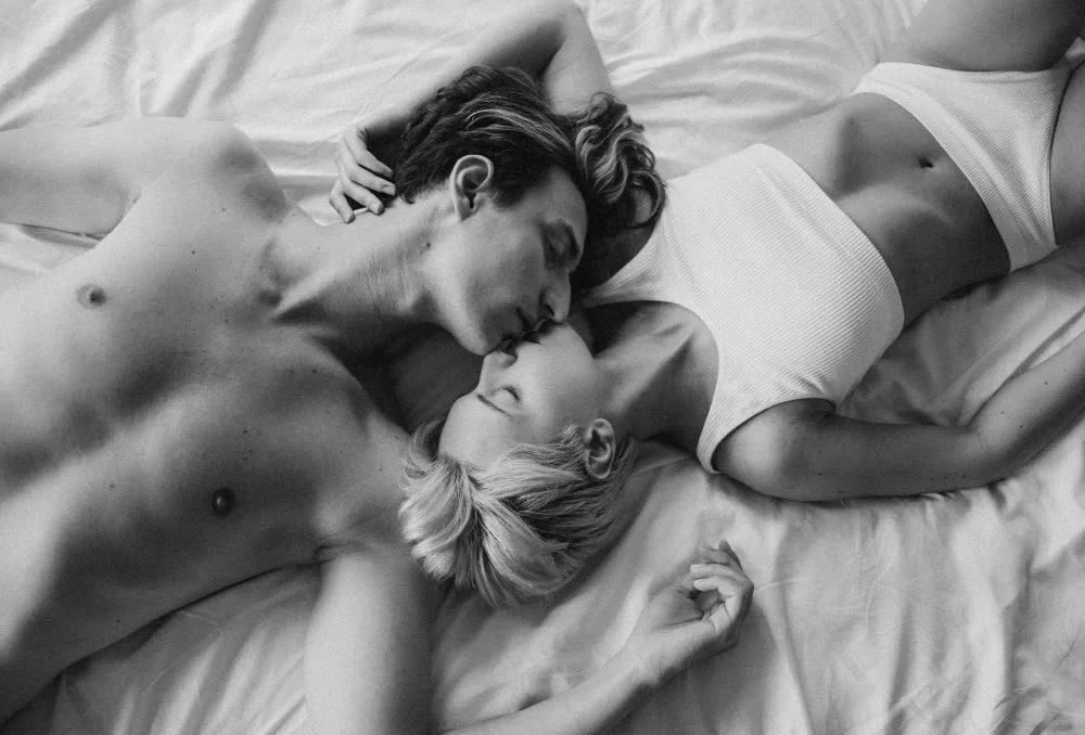 A black and white photo of a shirtless man and a woman wearing a white tank top lying on a bed, facing each other and about to kiss.