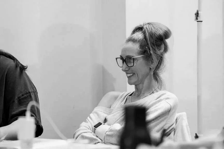 A woman with glasses, her hair in a bun, smiling with her arms crossed, seated at a table in a casual setting.