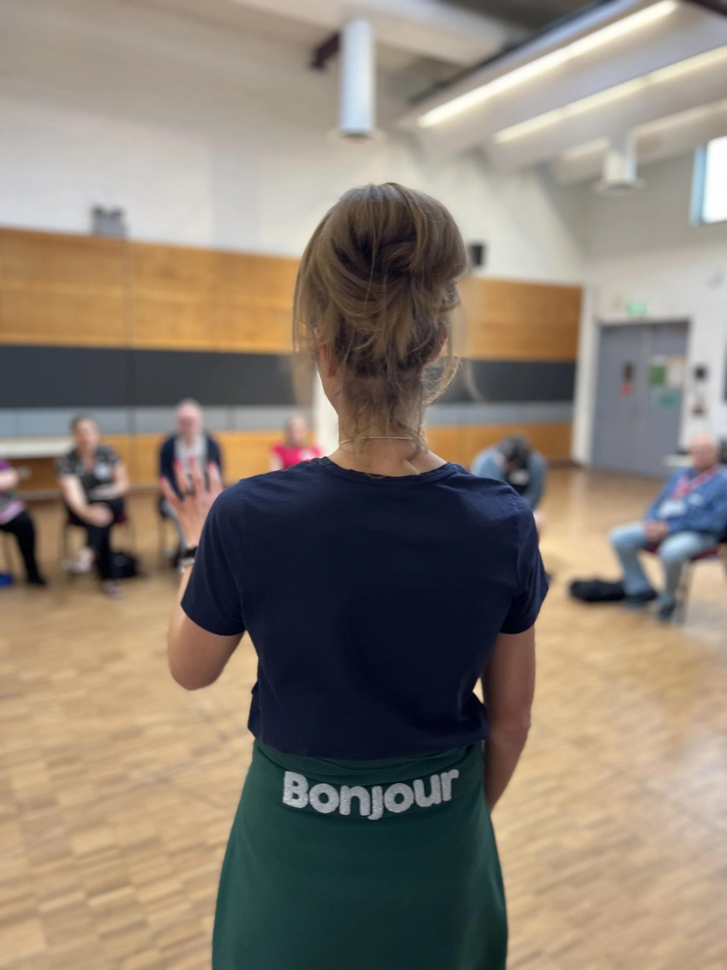 A woman with brown hair in a bun facing a group of people sitting in chairs in a room with wood floors and white walls.