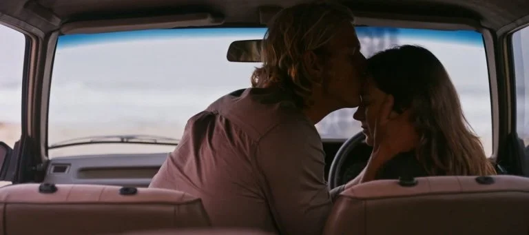 A man and a woman sitting in the front seats of a vehicle, sharing a kiss, with outside scenery visible through the windshield.