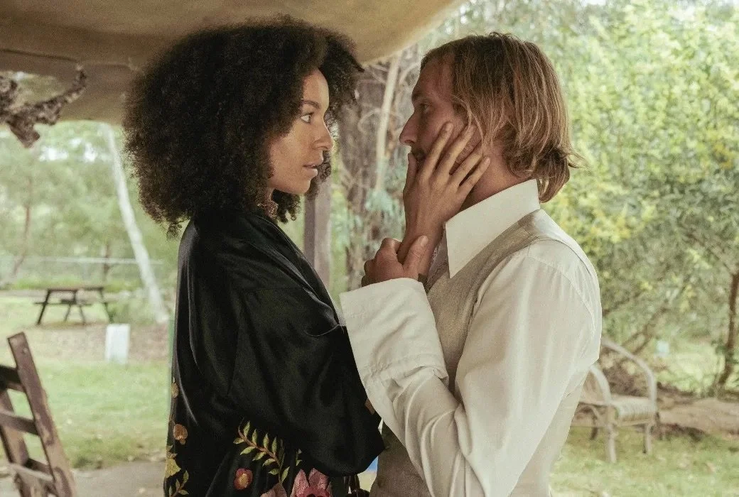 A woman with curly hair and a man with long blonde hair holding each other's faces and staring into each other's eyes in an outdoor setting.