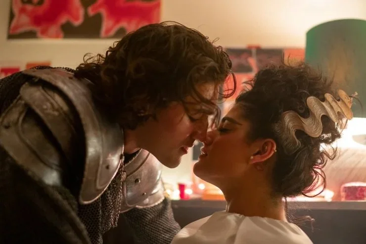 A romantic scene with a man in medieval armor leaning close to a woman with dark curly hair in an updo, touching noses in an intimate moment.