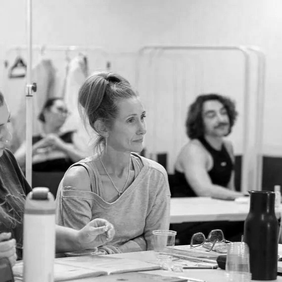 A woman with a high ponytail sitting at a table, appearing focused, with other people and a transparent partition in the background.