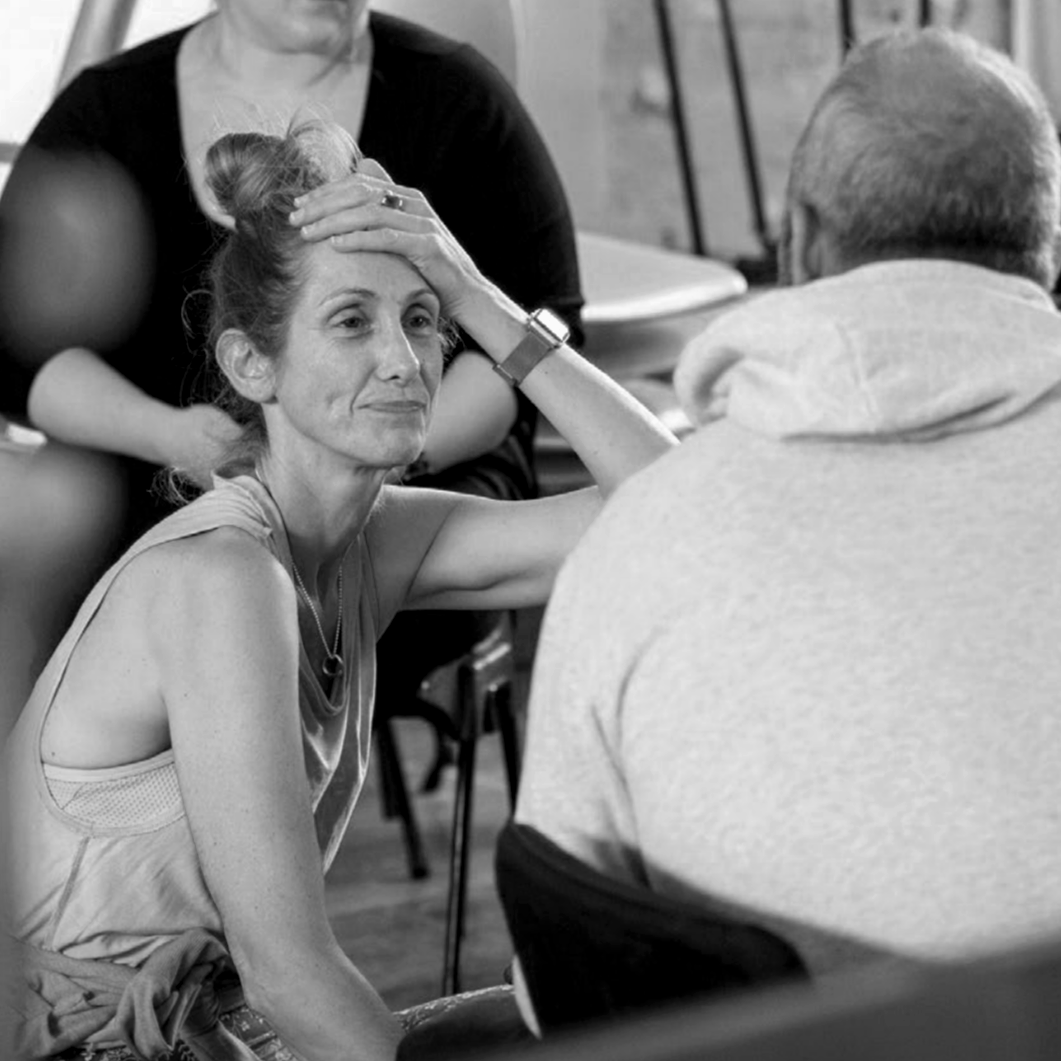 A woman with an emotional expression, sitting with her hand on her forehead, while talking to a man whose back is turned to the camera. Another person stands behind her, with their hand on her head, comforting her.