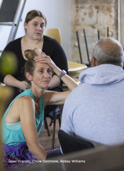 Three people in a casual setting, one sitting on the floor with her hair in a bun, another sitting with her hand on her head, and a third woman standing behind them, all engaged in conversation.