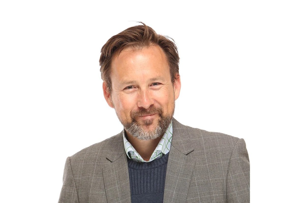 A middle-aged man with brown hair, a beard, wearing a brown checkered blazer, a green and white plaid shirt, and a navy sweater, smiling slightly and looking at the camera.