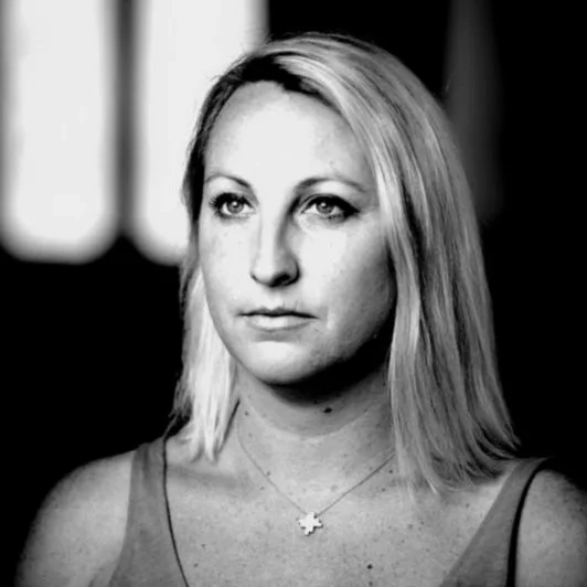 Black and white portrait of a woman with shoulder-length blonde hair, wearing a sleeveless top and a necklace with a cross pendant, looking to the side.