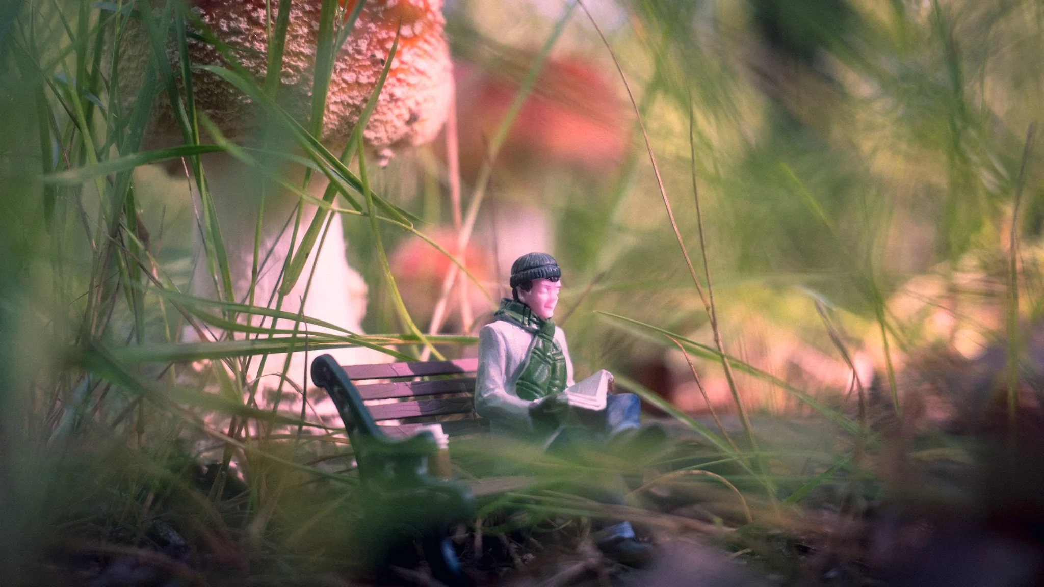 A miniature toy figure of a man sitting on a park bench, reading a newspaper, surrounded by tall grass and mushrooms.