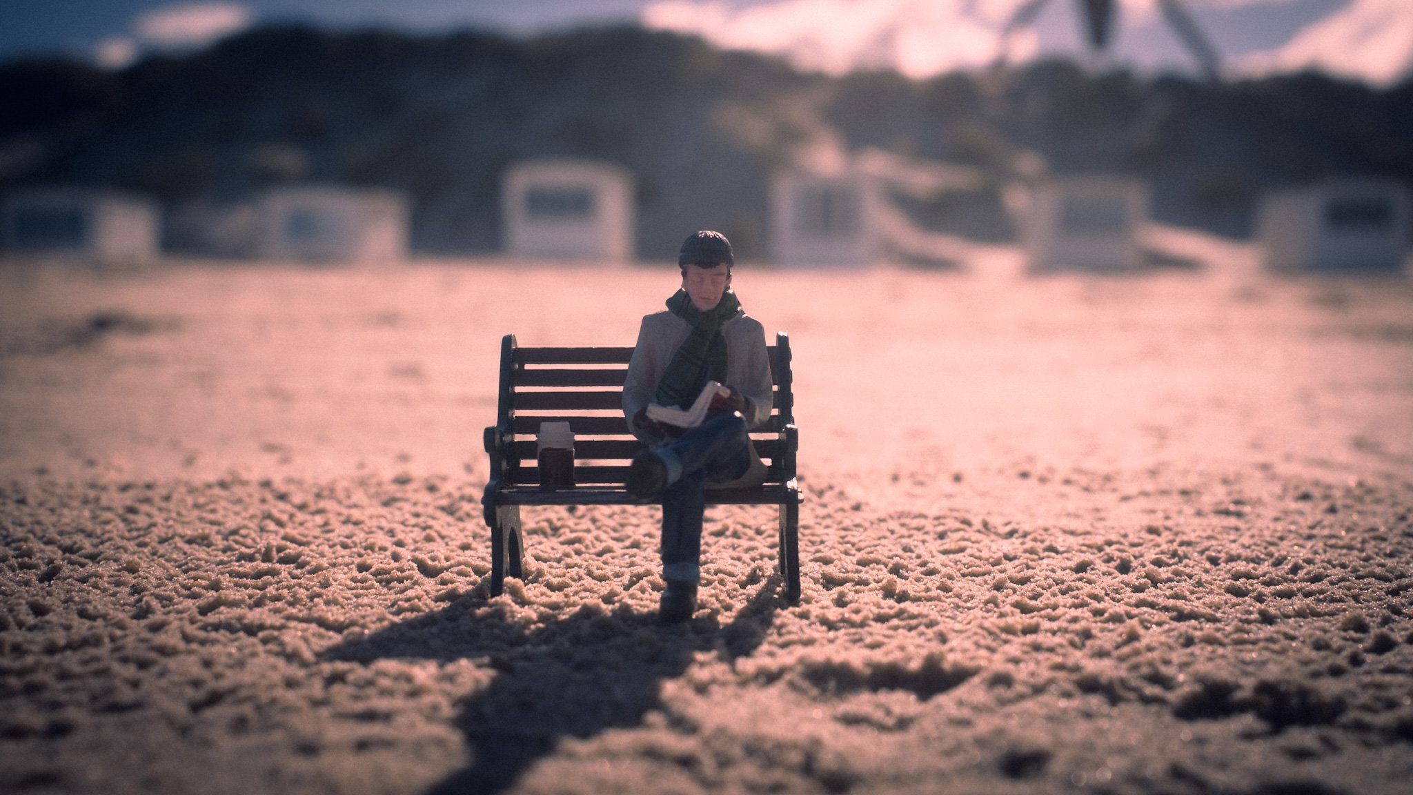 A person sitting on a bench on a sandy beach at sunset, reading a book, wearing a hat and scarf.