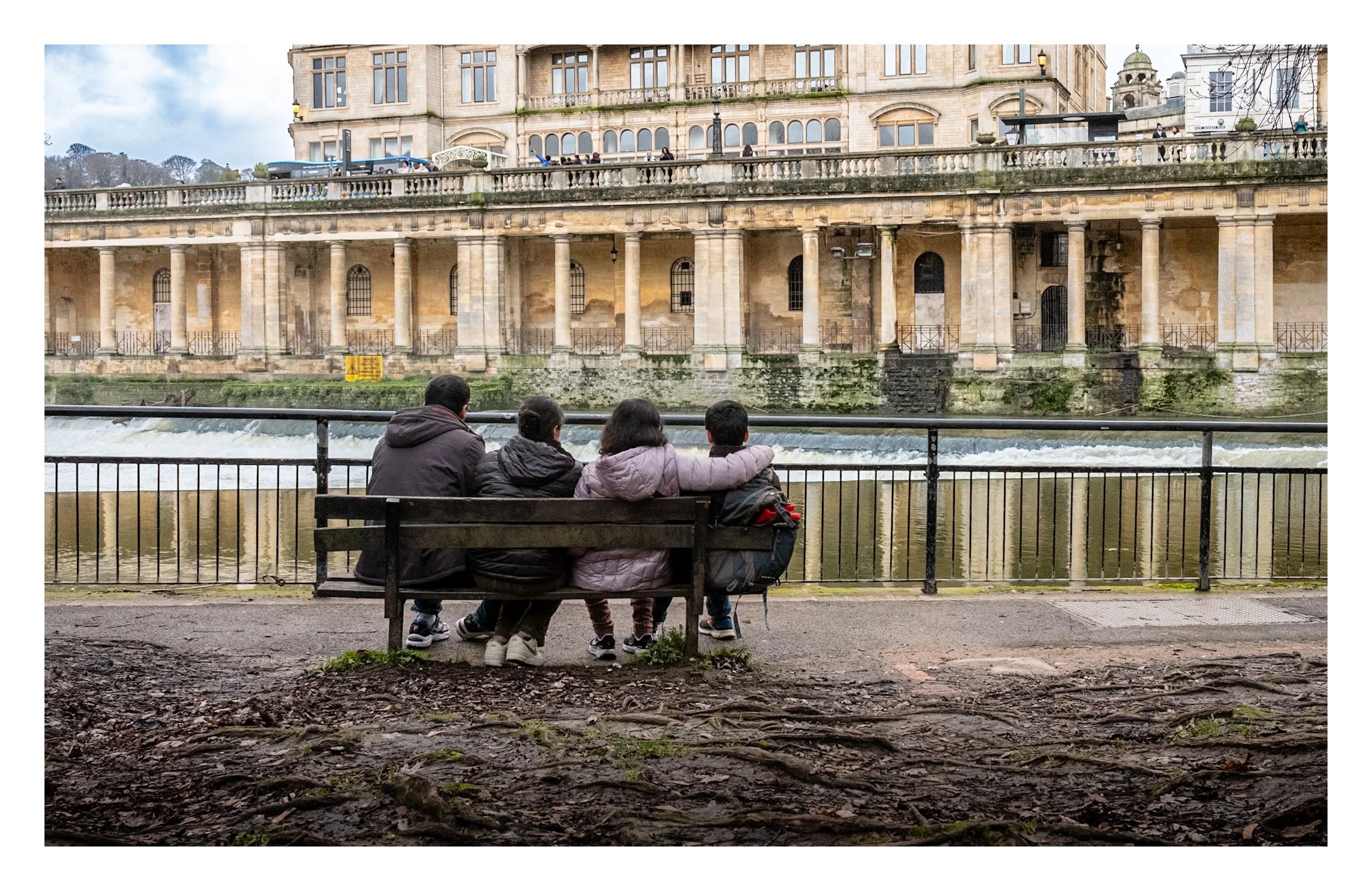 Four people sitting on a bench near a river, with historic stone buildings in the background, in an urban park setting.