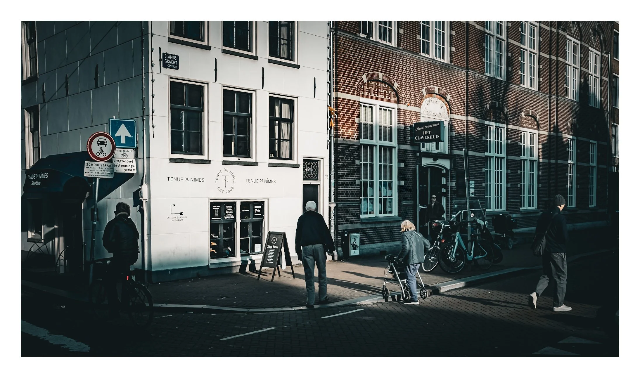Street scene in Amsterdam with pedestrians, bicycles, and old brick and white buildings. Shadows cast by buildings and people on the pavement.