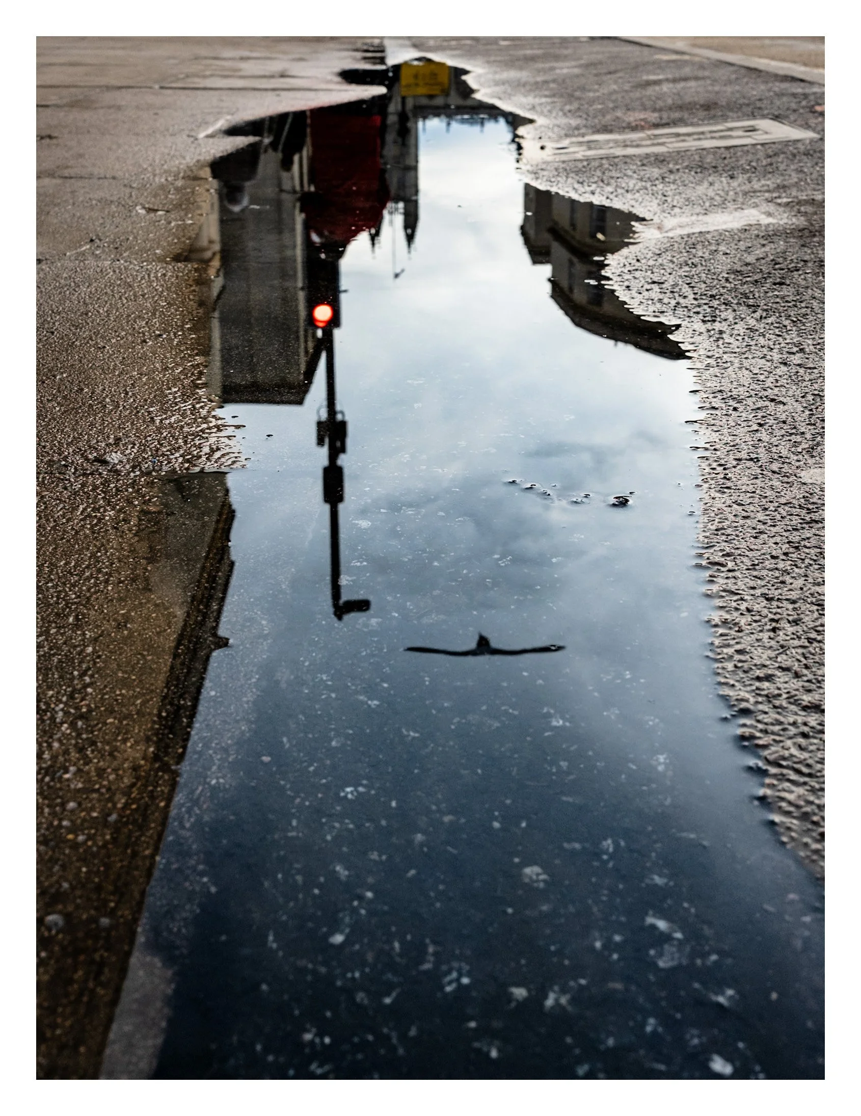 Reflection of a building, traffic light, and cloudy sky in a puddle on a city street.