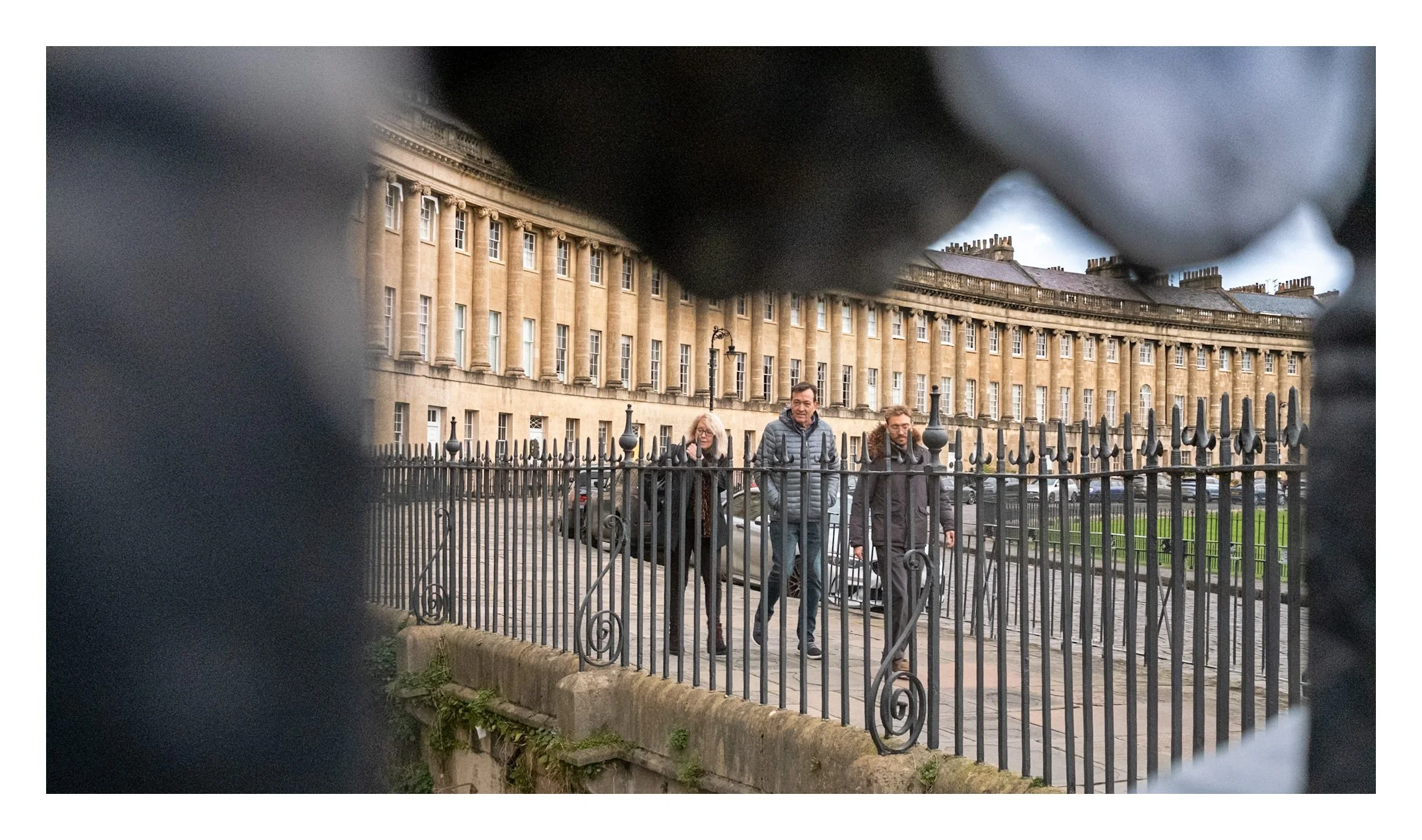 View through a black iron fence of three people walking past a historic building with yellow stone facade and multiple windows in an urban setting.
